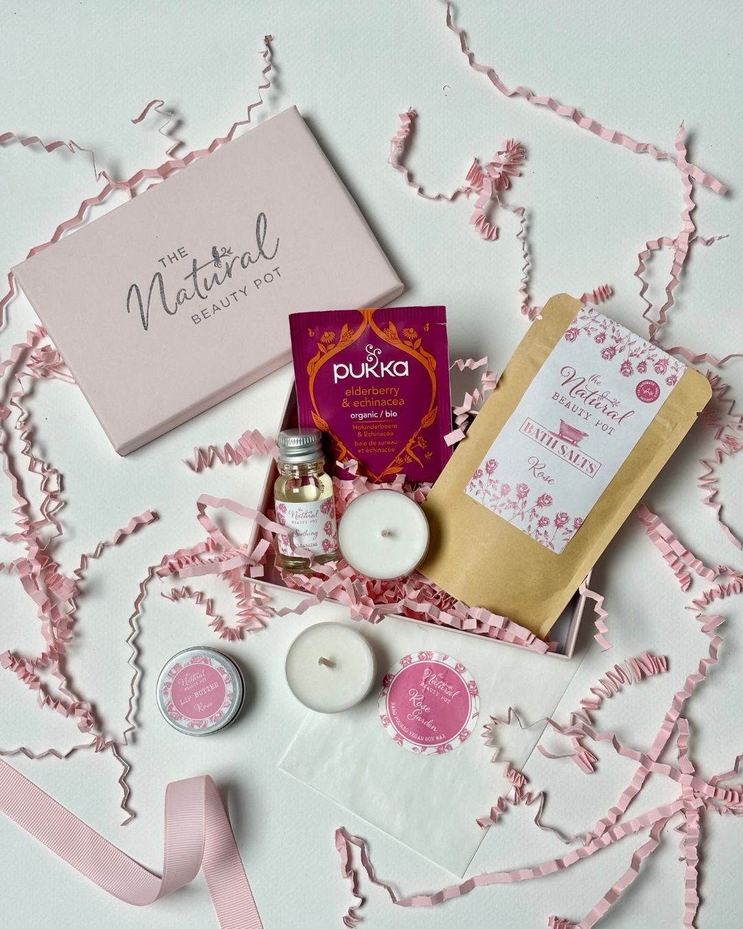Collection of natural beauty products including tea light candles, tea, bath salts, lip balm and skincare items on a light background with decorative ribbons.