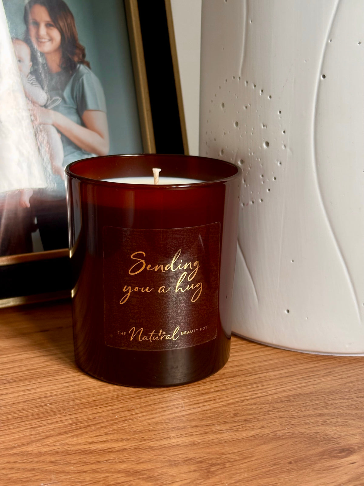 A dark amber glass jar candle with a wick and a printed label reading 'Sending you a hug' and 'the Natural Scents Co.' on a wooden surface, with a framed photo and a white textured vase in the background.