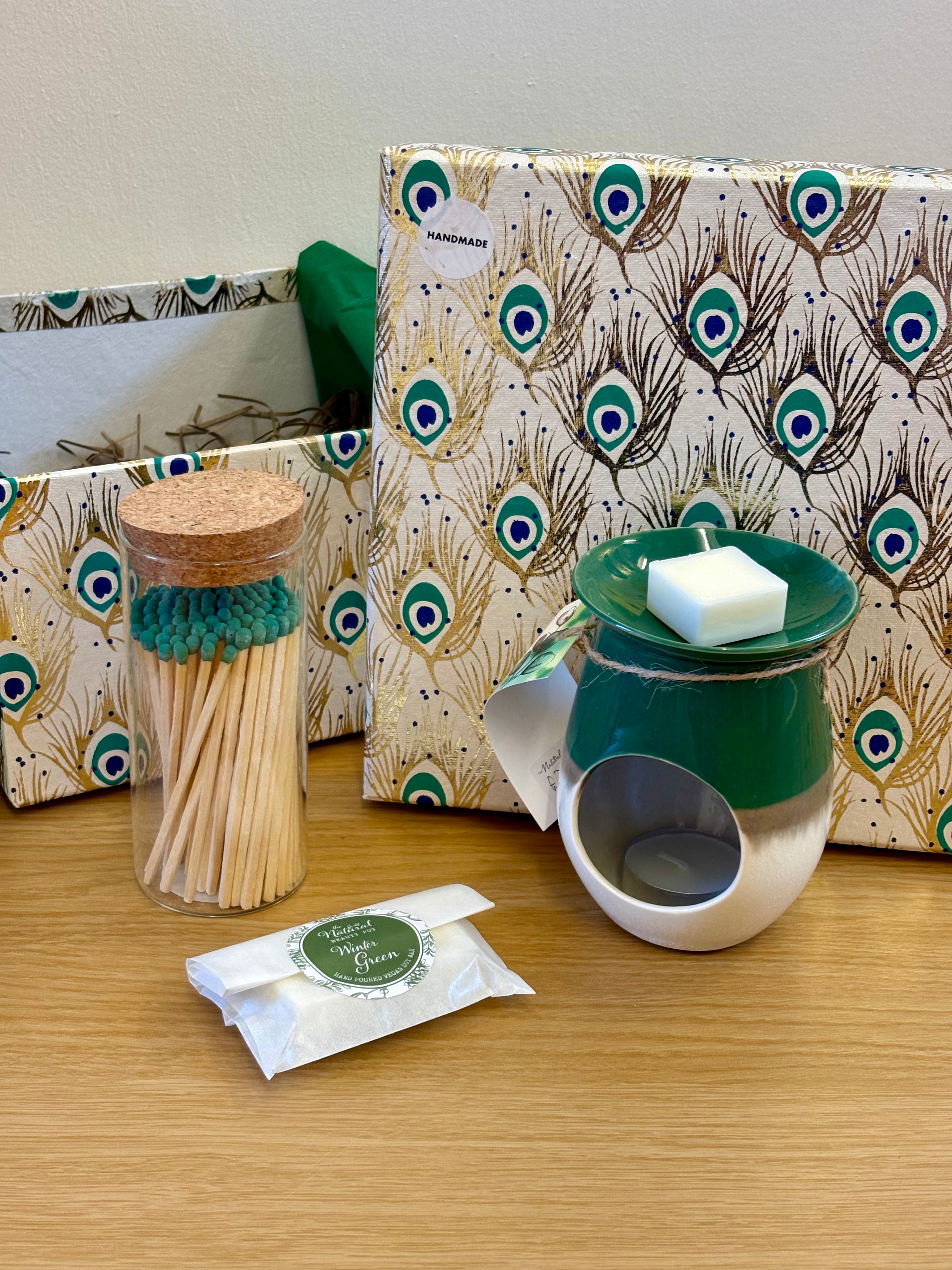 A gift set on a wooden table featuring a green ceramic wax warmer with a white candle, a glass jar of coloured  matches a wax melts by the Natural Beauty Pot matching gift boxes with a peacock feather pattern.