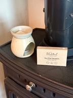 A box of 'The Natural Beauty Pot' 'Bu the hearth' wax melts sits next to a speckled cream ceramic wax warmer on a dark wood stove.