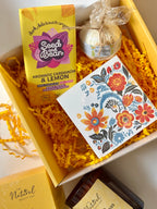 Gift box with 'Seed and Bean' cardamom and lemon chocolate, a bath bomb, and a floral matches and candle on yellow gift box.