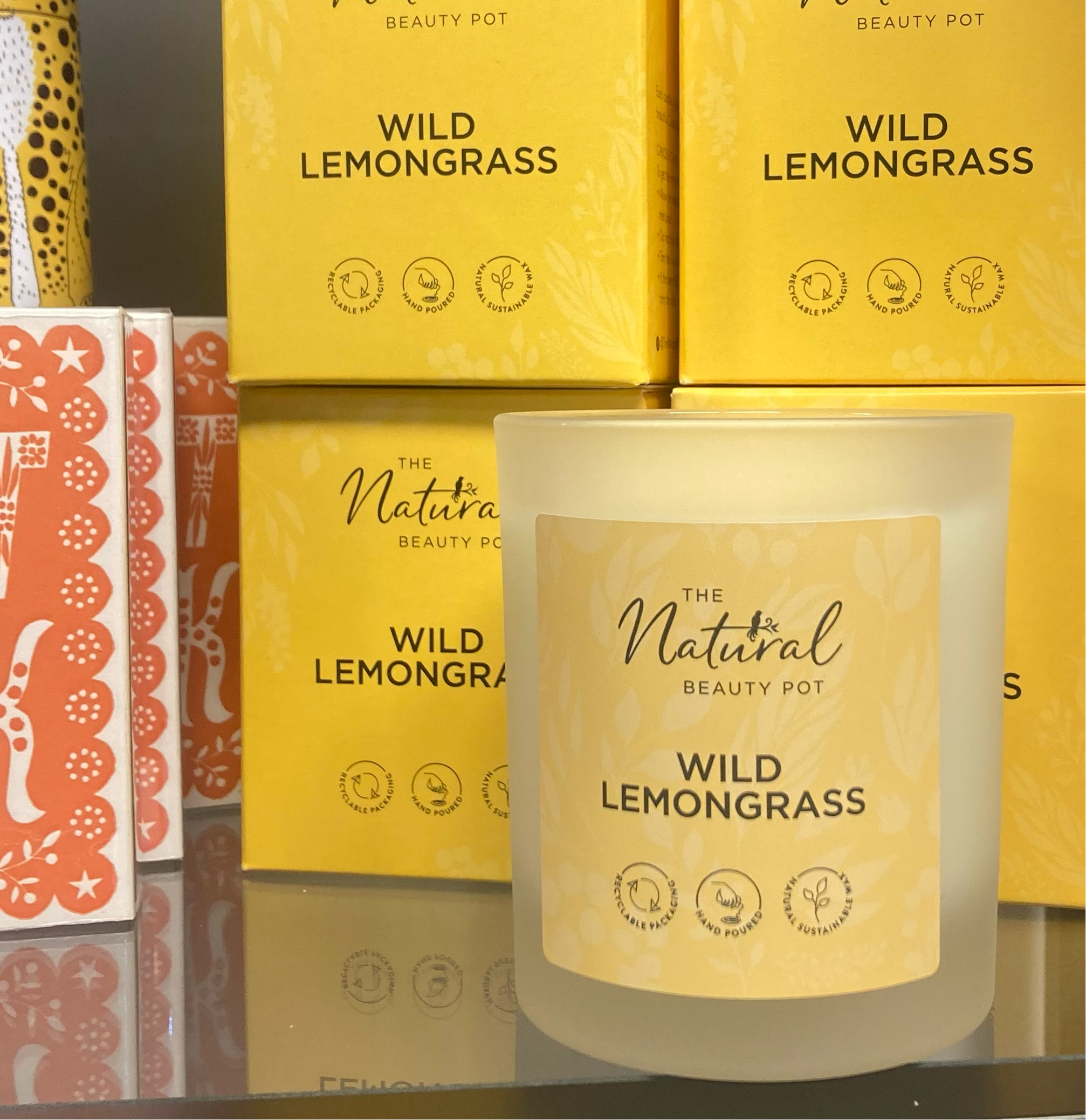 A clear soy candle with a label reading 'Wild Lemongrass, The Natural Beauty Pot,'. The candle is displayed in front of a yellow box with patterned design and the brand's name on it.