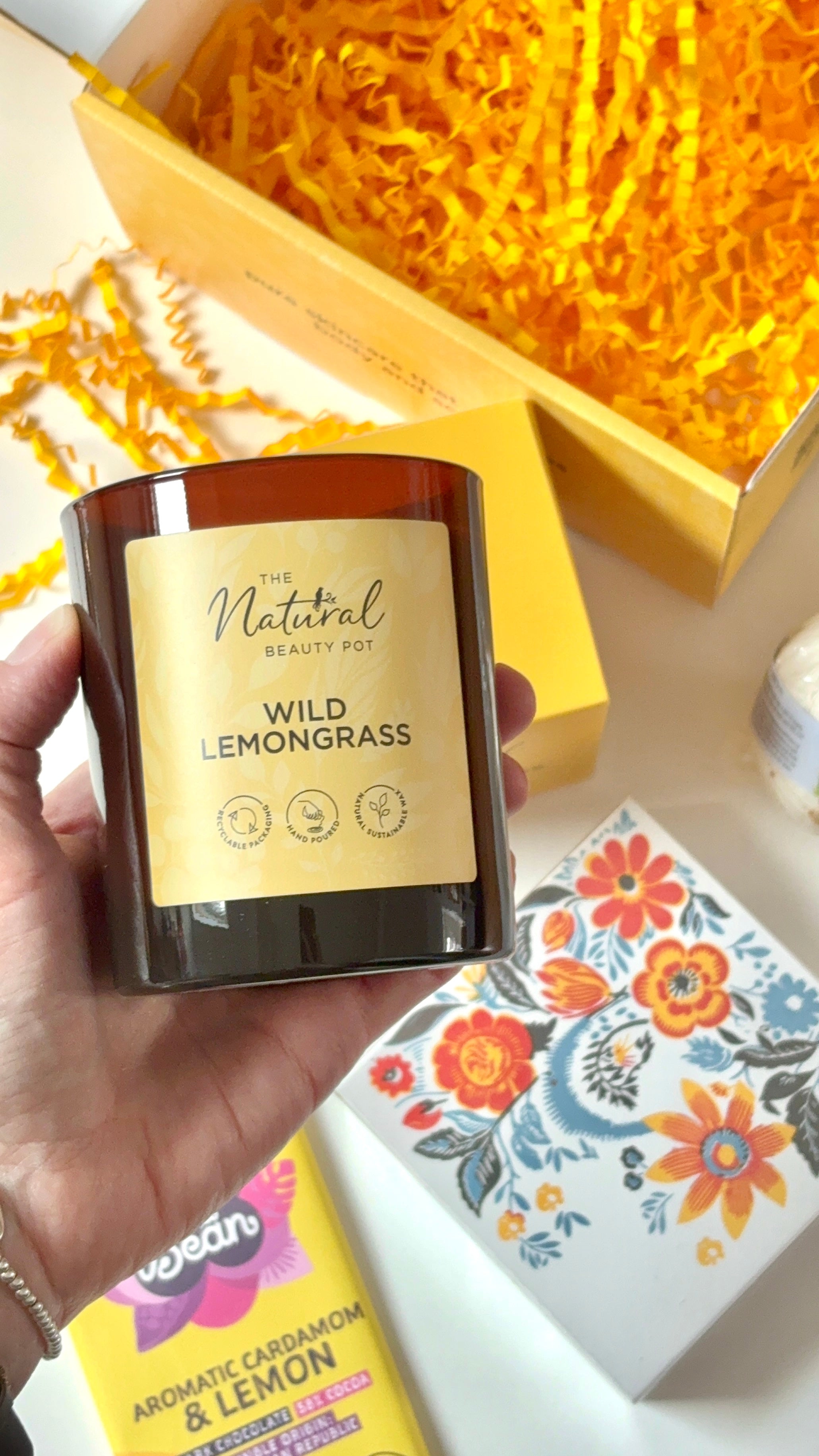 Hand holding a candle labeled 'Wild Lemongrass' with a decorative box and floral notebook in the background.