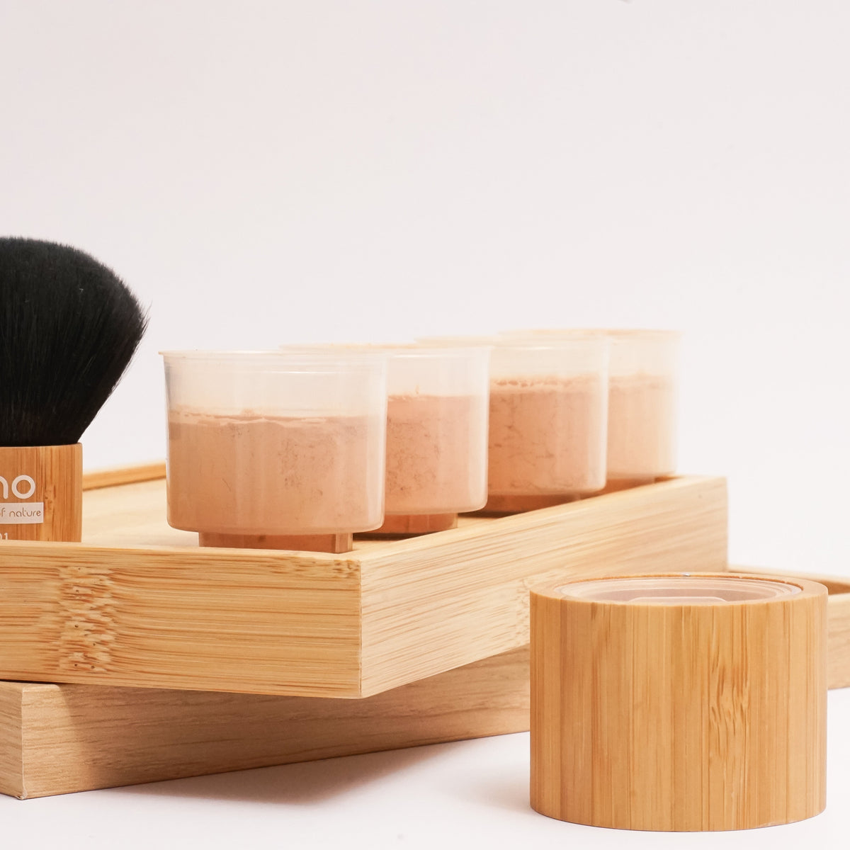 ZAO makeup set in bamboo, featuring a black-handled brush, four small clear jars filled with mineral silk foundation, and a round wooden pots, arranged on a light wood tray against a white background.