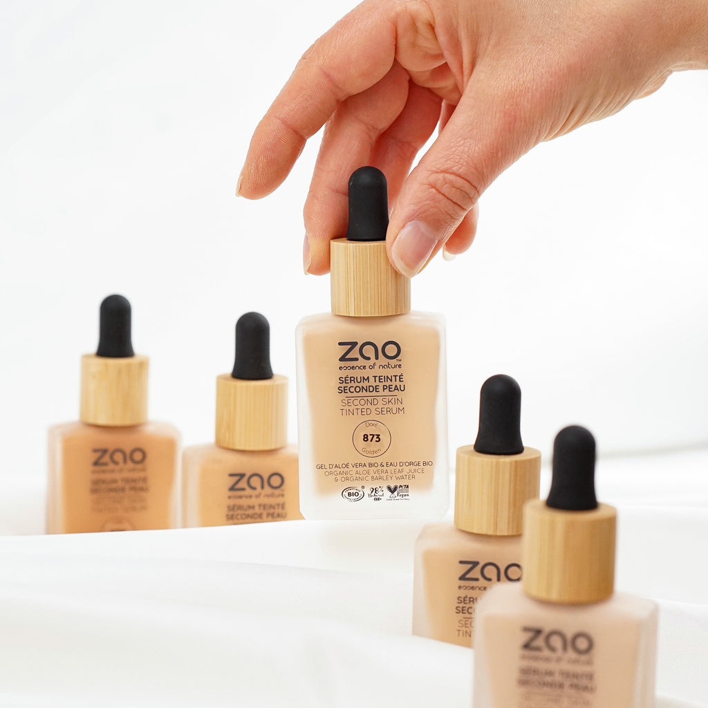 A hand holds a bottle of ZAO Second Skin Serum Tint in shade R5, with four other bottles of the same product arranged on a white draped fabric surface against a white background.