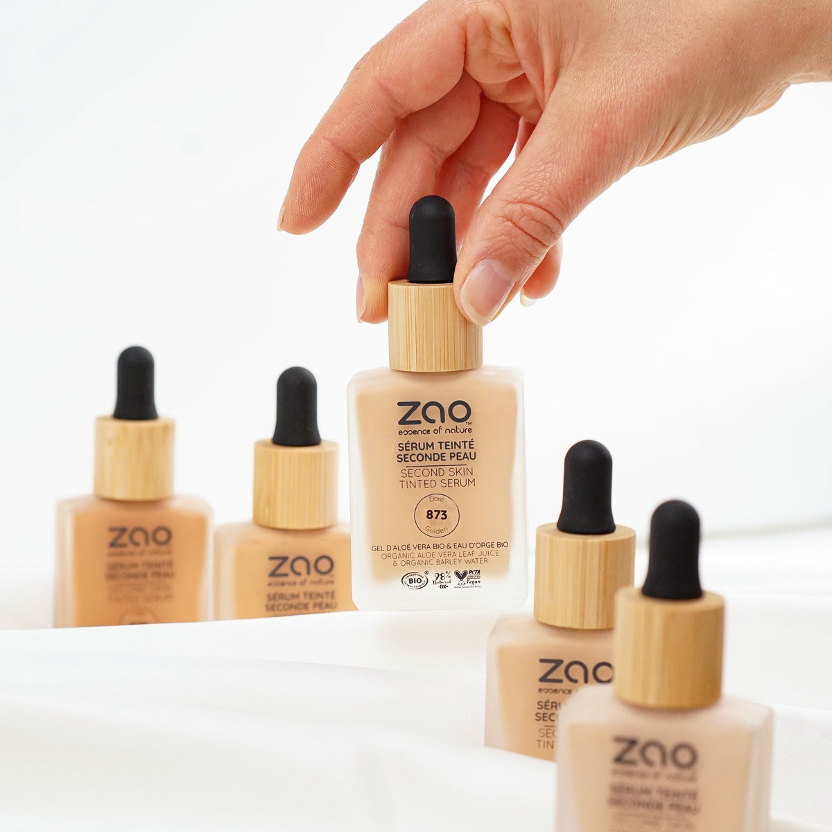 A hand holds a bottle of ZAO Second Skin Serum Tint in shade R5, with four other bottles of the same product arranged on a white draped fabric surface against a white background.