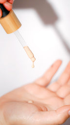 A close-up of a clear dropper with beige liquid foundation being held above an open hand, showing the product being dispensed.