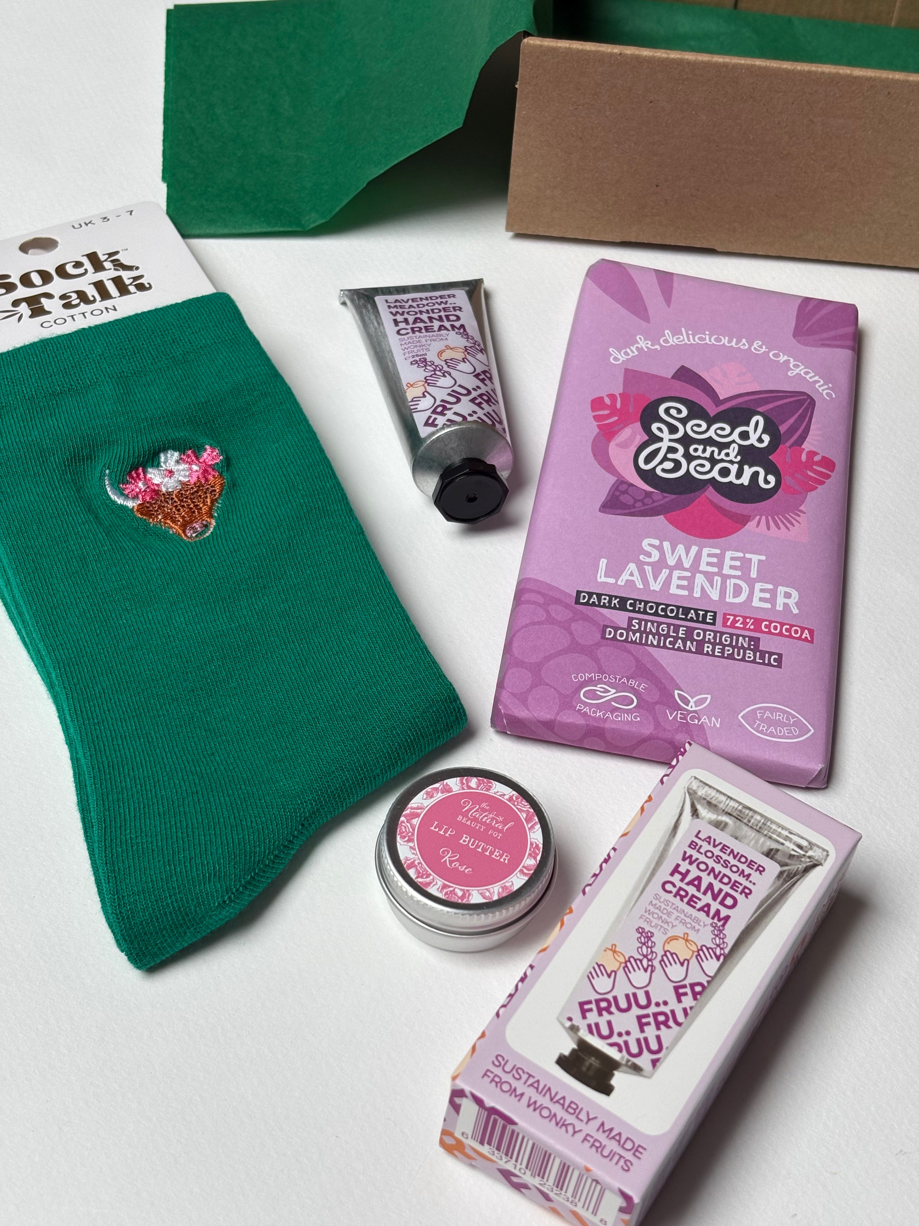 A green, pink and purple gift set arranged including  green bamboo socks, a purple bar of Seed and Bean Sweet Lavender chocolate, a lavender hand cream tube, rose lip butter, alongside green tissue paper and a cardboard gift box.
