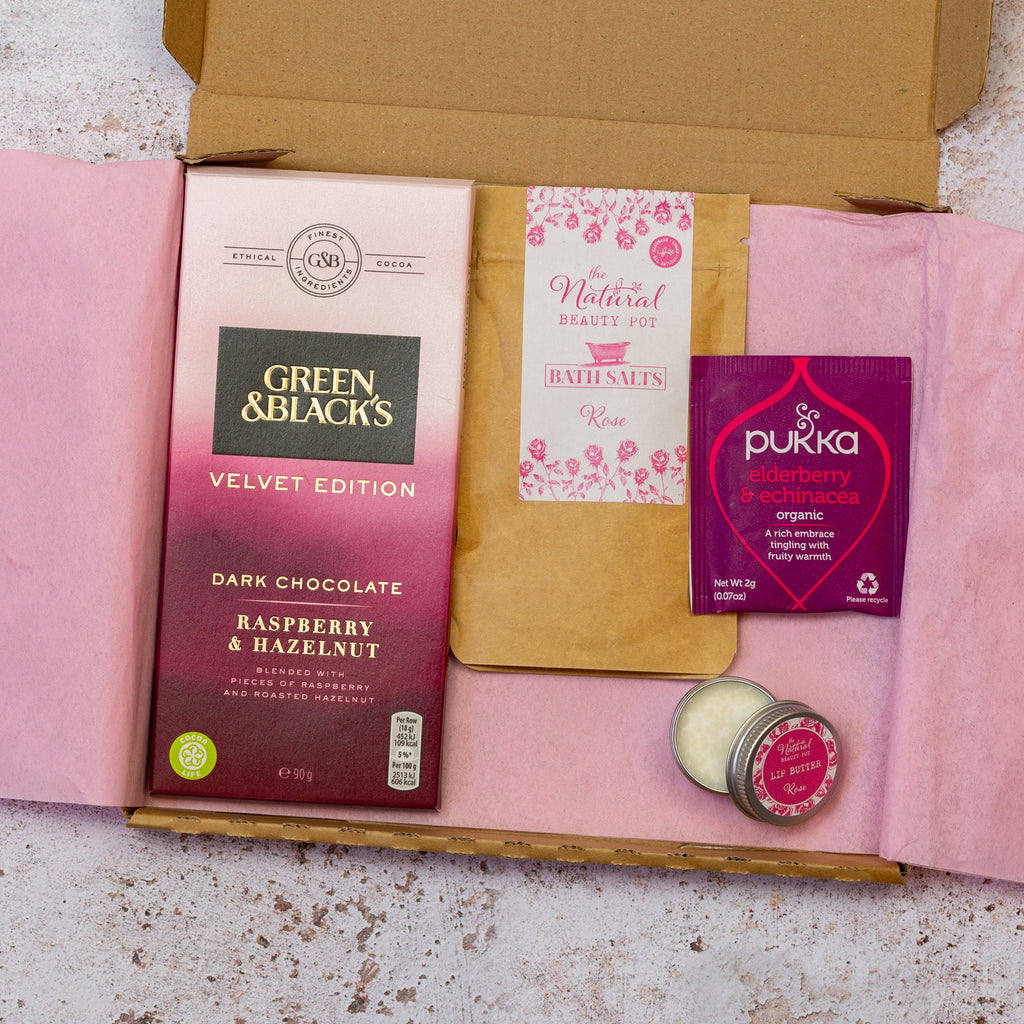 A gift set containing a vegan chocolate bar, a pink sachet of rose bath salts, a herbal tea bag, and a small open lip balm arranged in a pink letterbox-style packaging.