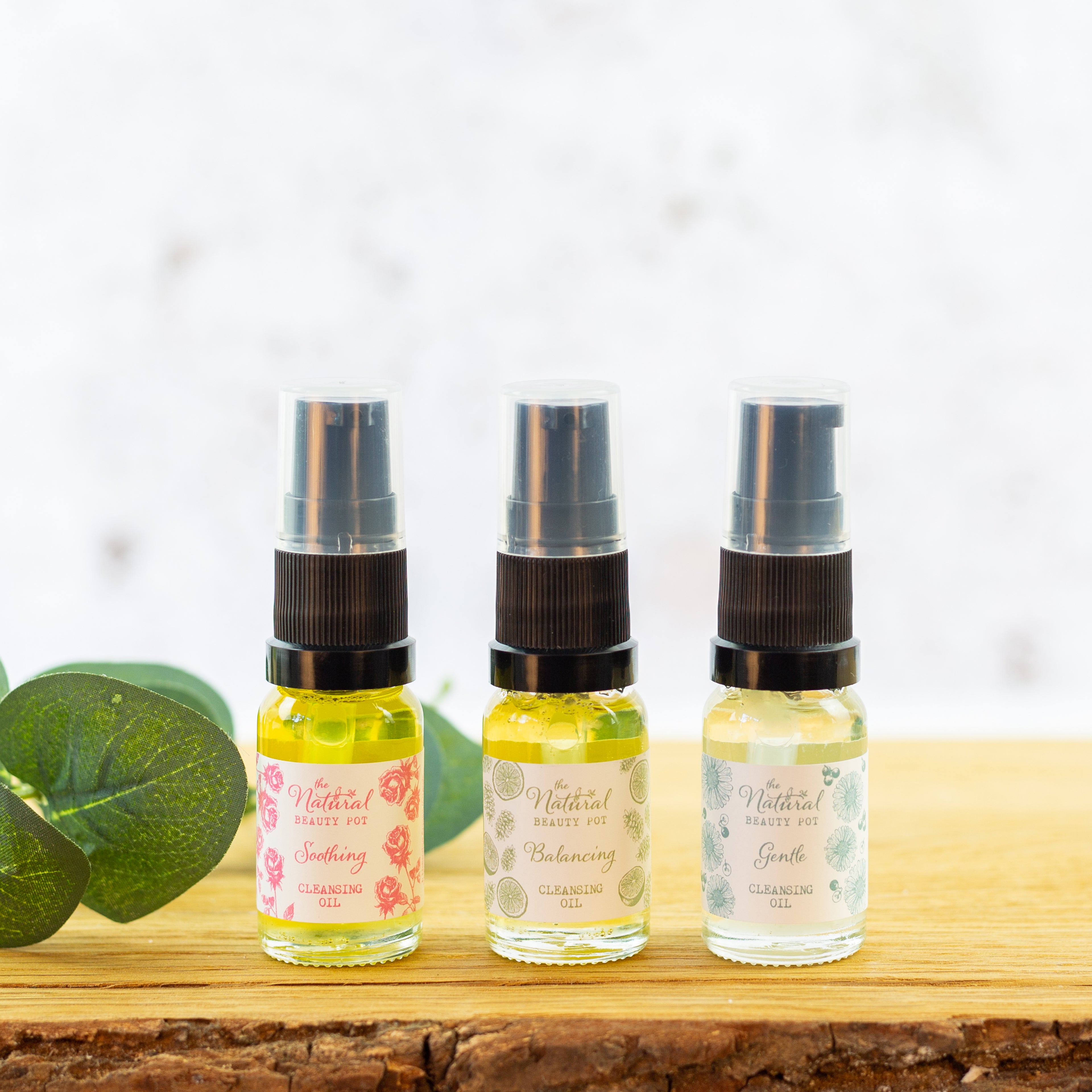 Three travel size bottles of cleansing oil with pump dispenser, each with different labels indicating the scent or type of oil. The bottles are set on a wooden surface with a green leafy background.