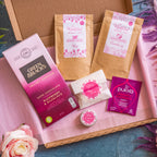 A letterbox gift set with various self-care products including a rose lip butter, bath salts, herbal tea, facial grains mask, and chocolate.