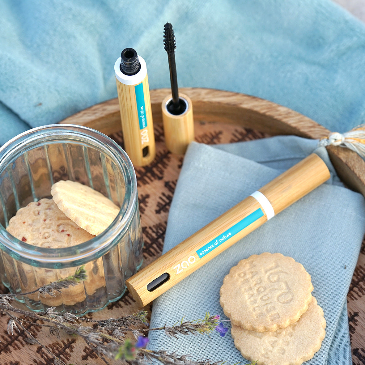 The image shows a tube of Zao Definition Mascara with its cap and wand visible, alongside a bamboo packaging container. There are also two cookies and a jar on a cloth surface.